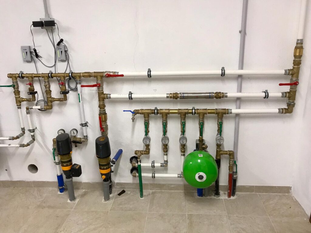 Installierte Heizungs- und Wasserverteilungseinheit mit Messgeräten, Ventilen, Pumpen und farbcodierten Rohren an weißer Wand.