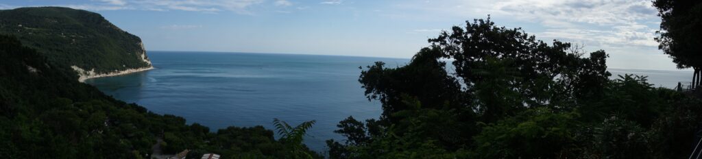 Weitläufige Küstenlandschaft mit bewaldetem Hügel, ruhiger Bucht und blauem Meer unter Wolkenhimmel.