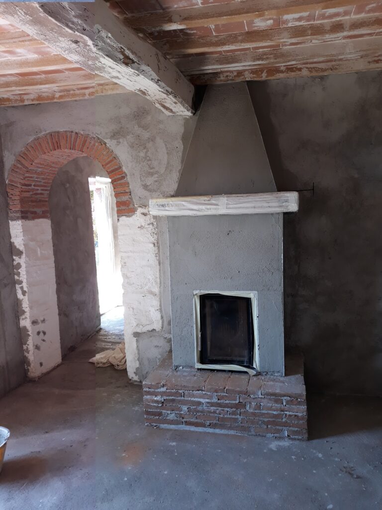 Unfertiger Innenraum mit gemauertem Kamin, weißem Holzrahmen, Ziegelbogen und freiliegenden Deckenbalken.