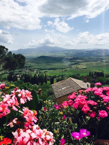 Mediterrane Landschaft mit blühendem Garten, rustikalem Steinhaus, Zypressen und Hügeln in der Toskana bei Sonnenlicht.